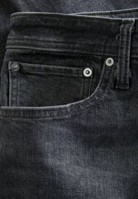 Lomme på sorte denimjeans med sømme og metalnitter, der viser bæltestrop og detalje af det teksturerede stof.