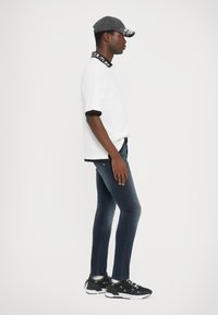 Λευκό μπλουζάκι με μαύρο γιακά και μανίκια, σκουρόχρωμο στενό τζιν και μαύρα αθλητικά παπούτσια με λευκές λεπτομέρειες και κορδόνια. Γκρι καπέλο με κείμενο.