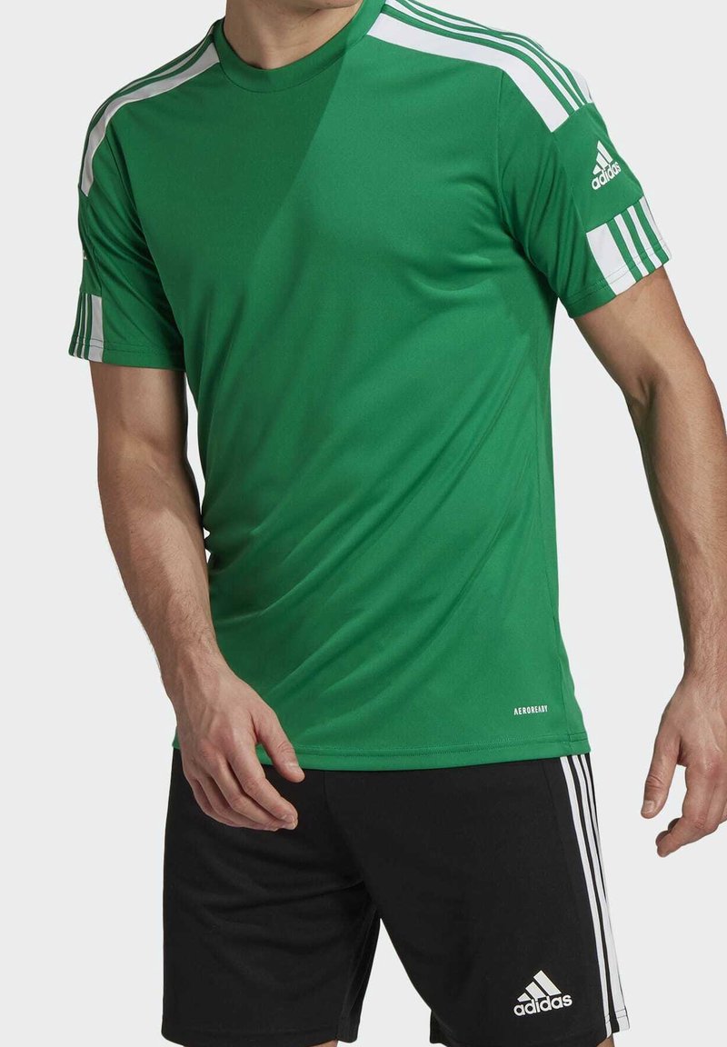 Man i en grön Adidas sporttröja med vita ränder på ärmarna och svarta Adidas shorts med vita ränder på sidorna.