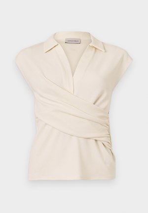 Kremfarget ermeløs omslagstopp med krage og sidesammensyinger, merket Anna Field, vist på ensfarget bakgrunn.