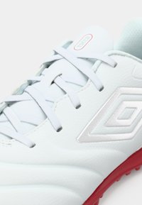 Zapatilla de fútbol blanca con parte superior sintética texturizada, cordones grises, suela roja y acento de logo plateado. Vista en primer plano de los detalles del diseño.