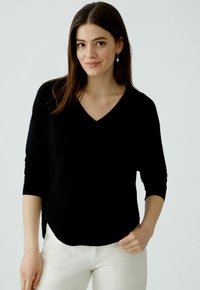 Schwarzer V-Ausschnitt-Pullover aus weichem Material, mit Dreiviertelärmeln und abgerundetem Saum. Kombiniert mit hellen Hosen.