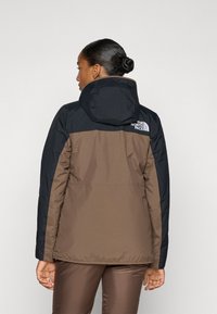 Hnedo-čierna kapucňová bunda s prešívaným dizajnom. Má logo The North Face na chrbte a hladkú textúru, vhodná na chladné počasie.