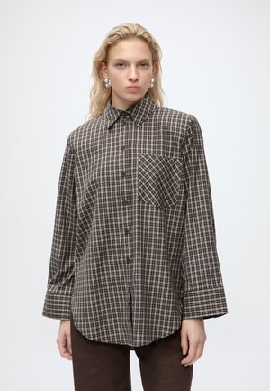 Femme aux cheveux blonds mi-longs portant de grandes boucles d’oreilles argentées en forme de créoles et une chemise ample à carreaux marron avec un pantalon foncé.