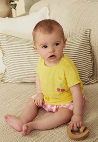 T-shirt jaune avec un détail de poche en cerise, à manches courtes, associée à un short rayé rose et blanc ; assis sur une surface beige texturée.