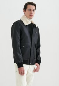 Svart läderjacka med en vit pälsartad krage, blixtlås, två framfickor och ribbade ärmslut. Slät textur och figursydd design.