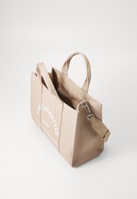 Beige lærred tote taske med dobbelte håndtag, der har en lynlås-lukning og hvidt trykt branding. Bemærkelsesværdigt hardware inkluderer en aftagelig rem.