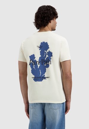 Man met krullend donker haar draagt een crèmekleurig T-shirt met een blauwe cactusafbeelding en de tekst "Pure Path" en "Own Your Journey" op de achterkant, gecombineerd met lichte spijkerbroek.