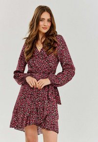 Kvetinové šaty v ružovej a čiernej farbe s dlhými rukávmi, zavinovacím strihom a volánovým lemom. Obsahujú malé biele a zelené kvetinové vzory po celom povrchu.