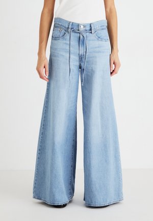 Persoon die lichtblauwe wijde jeans draagt met een trekkoord in de taille en een wit mouwloos topje, staand tegen een effen achtergrond.