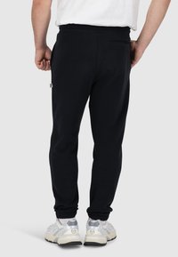 Zwarte joggers met een trekkoord in de taille, elastische boorden en een achterzak. Zacht materiaal met een gladde textuur. Neutrale sneakers worden gedragen.