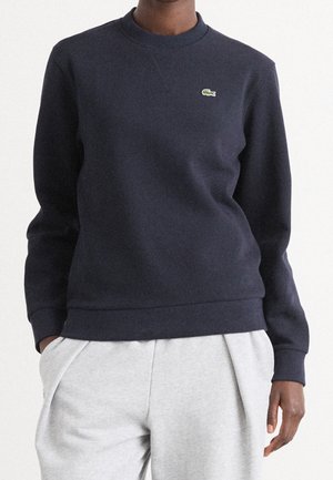 Personne portant un sweat-shirt bleu marine avec un petit logo crocodile vert sur la poitrine et un pantalon gris clair, mains dans les poches, sur fond uni.