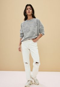 Felpa grigia con ricami floreali, abbinata a jeans bianchi strappati e sneakers grigie. Tessuto testurizzato, vestibilità ampia, design casual.