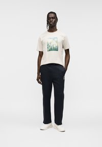 Ανοιχτό μπεζ μπλουζάκι με πράσινο γραφικό σχέδιο, συνδυασμένο με ναυτικό φόρμας με λογότυπο και λευκά αθλητικά παπούτσια.