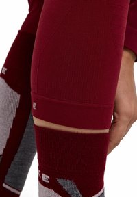 Close-up van een persoon die bordeauxrode textuurleggings en bijpassende sokken met grijze accenten draagt, hand rustend nabij de knie.