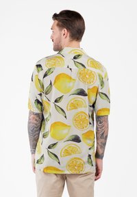 Hombre con brazos tatuados de espaldas, vistiendo una camisa de manga corta con un patrón de limones amarillos y hojas verdes y pantalones beige.