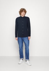 Camicia a maniche lunghe blu navy con chiusura a bottoni e tasca sul petto, abbinata a jeans azzurri chiari e sneakers bianche. Tessuto morbido e vestibilità casual.