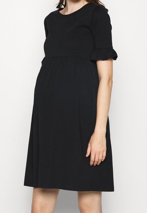 Femme enceinte portant une robe de maternité noire à manches courtes avec des manches à volants, debout devant un fond clair uni.