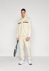Chándal deportivo color crema con acentos de rayas negras. Presenta una chaqueta con cremallera y capucha, puños elásticos y pantalones a juego. Sosteniendo una raqueta de tenis.