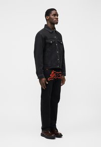 Μαύρο τζιν μπουφάν με κουμπιά, δύο τσέπες στο στήθος και κόκκινος κεντητός σχεδιασμός γάτας σε μαύρο παντελόνι, συνδυασμένο με καφέ μποτάκια με κορδόνια.