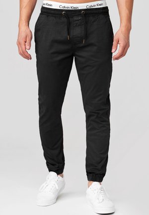 Man draagt zwarte joggerbroek met elastische boorden, witte Calvin Klein tailleband zichtbaar en witte sneakers tegen een effen achtergrond.
