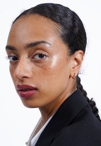 Porträt einer Person mit mittelbrauner Haut, Sommersprossen und einem seitlichen Nasenring. Sie trägt ein schwarzes Sakko über einem weißen Hemd und hat glänzende Lippen.