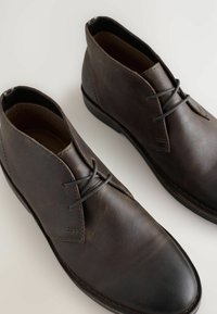 Botas chukka de cuero marrón con una textura suave, que presentan una punta redondeada, detalles de costura y cordones en contraste sobre una suela de perfil bajo.