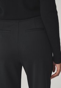 Svarta figurnära byxor med en slät yta, utrustade med midjehöjd och två diskreta bakfickor utan synliga detaljer.