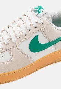 Sapatilha Nike Air Force 1 branca e bege com swoosh verde, atacadores brancos e sola de borracha de cor caramelo, apresentada em close-up de perfil.