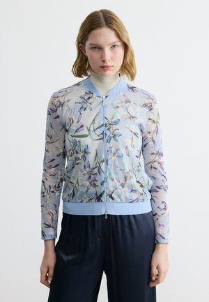 Femme portant une veste zippée bleu clair à motifs floraux sur un col roulé beige, associée à un pantalon noir, debout devant un fond blanc uni.