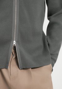 Grå hettegenser med glatt tekstur, med en metallglidelås og flate sømmer, båret over beige bukser med knappedetaljer.