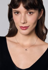 Srebrny naszyjnik z zawieszką w kształcie pszczoły, z delikatnymi detalami. Noszony przez osobę w bluzce z dekoltem w kształcie litery V. Gładka tekstura skóry, neutralne tło.