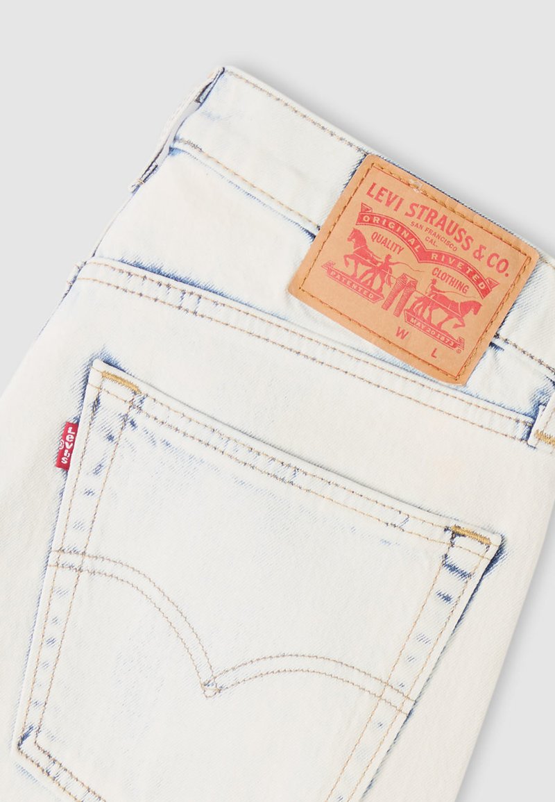 Jean en denim blanc avec une poche arrière cousue, un patch en cuir marron avec logo, et un détail de languette rouge sur la couture latérale.