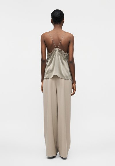 Γυναίκα που στέκεται με την πλάτη προς το μέρος μας, φορώντας αμάνικη σατέν μπλούζα με δαντελένιο τελείωμα και λεπτές τιράντες, καθώς και μπεζ παντελόνι με φαρδιά μπάσκα.