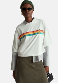 Biała krótka koszulka z tęczowymi paskami, z grafiką żyrafy; nałożona na pasiastą bluzkę z długim rękawem z zielonymi elementami.