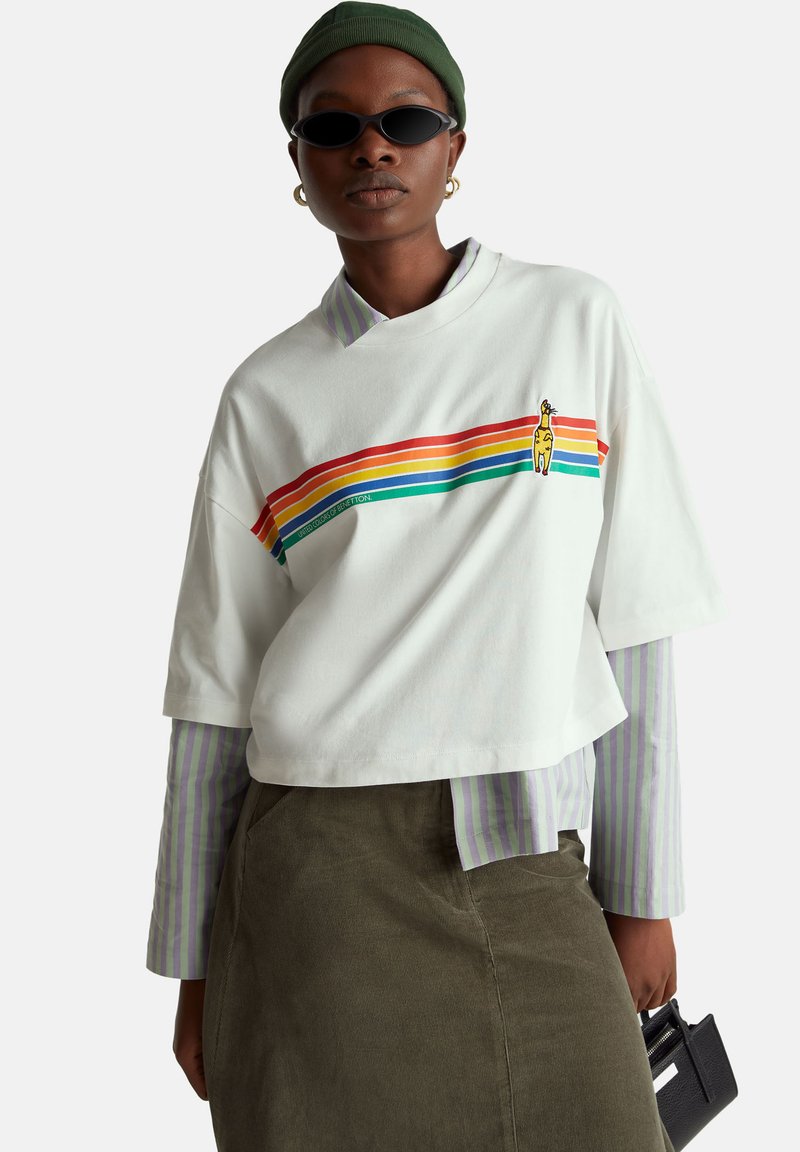 Biała krótka koszulka z tęczowymi paskami, z grafiką żyrafy; nałożona na pasiastą bluzkę z długim rękawem z zielonymi elementami.