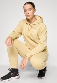 Beige hoodie med framficka och emblem, kombinerad med matchande tajta leggings och svarta löparskor med rosa detaljer.
