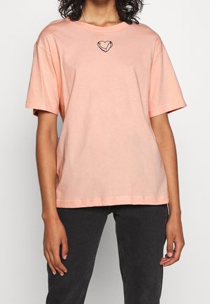 Frau trägt ein lockeres pfirsichfarbenes T-Shirt mit einem kleinen schwarzen Herzumriss auf der Brust, kombiniert mit schwarzen Hosen.