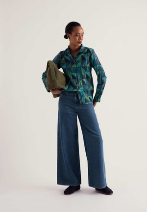 Vrouw staat met een olijfgroene tas, draagt een geometrisch patroon shirt, wijde spijkerbroek en donkere schoenen tegen een effen achtergrond.