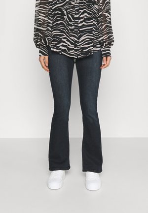 Person iført mørkeblå bootcut-jeans, zebramønstret bluse med lange ærmer og hvide sneakers, stående på en ensfarvet lysegrå gulv.