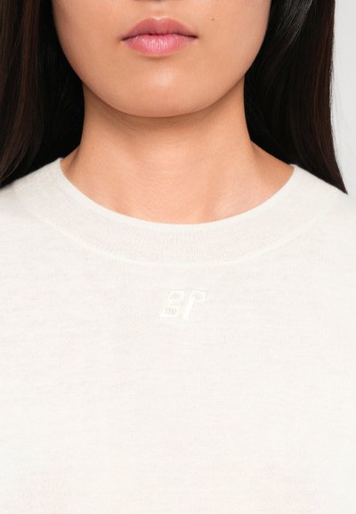 Kerge beež sviiter ümmarguse kaelusega, valmistatud pehmest kangast, millel on rinnaosas peen valge logo. Sile tekstuur.