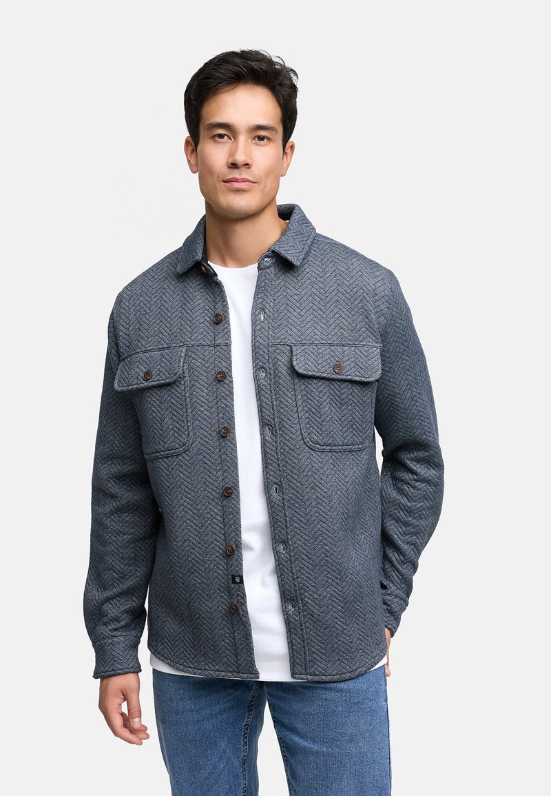 Giacca camicia a motivo chevron grigio con due tasche sul petto, chiusura a bottoni e colletto, indossata sopra una camicia bianca.