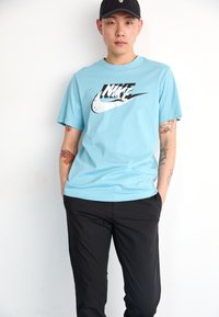 Light blue short-sleeve t-shirt featuring a black and white Nike logo on the front, paired with black pants. Smooth fabric texture, relaxed fit.