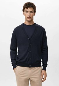 Granatowy kardigan z dekoltem w serek wykonany z miękkiego materiału, z przodu zapinany na guziki i wykończony prążkowanymi ściągaczami na rękawach. Nosi się go na ciemnym t-shircie i zestawia z jasnymi spodniami.