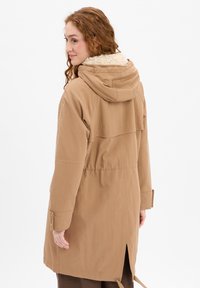 Knee-length tan coat with a hood, featuring a soft, fluffy collar, side pockets, and adjustable waist and cuffs. Smooth fabric texture.