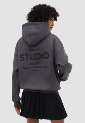 Vrouw met krullend haar draagt een oversized donkergrijze hoodie met zwarte tekst op de achterkant en een zwarte geplooide rok, zijwaarts gekeerd.