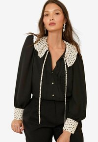 Donna che indossa un blusa nera con un ampio colletto e polsini a motivo cuori bianchi, maniche lunghe, bottoni a forma di cuore e pantaloni neri abbinati.