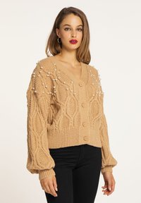 Cardigan tricotat bej, cu un decolteu în V adânc, model texturat și aplicații de perle. Include trei butoane și mâneci bufante.