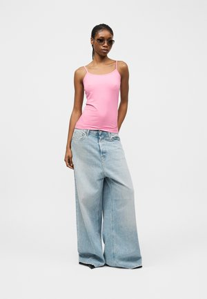 Kvinde iført solbriller, lyserød top med spaghetti-stropper og lyseblå jeans med vide ben, stående mod en ensartet lysegrå baggrund.