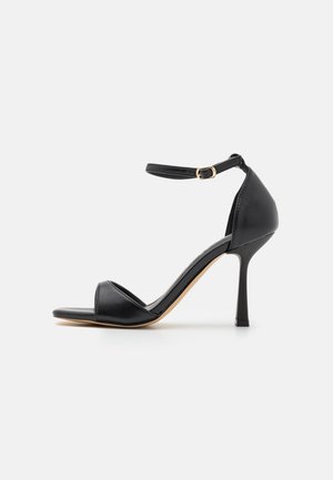 Sort højhælet sandal med en slank ankelrem, åben tå og blank overflade. Har en lige stilettohæl og beige sål.